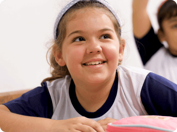 Menina sorridente sentada em uma carteira escolar, usando uniforme, com um estojo rosa sobre a mesa; ao fundo, outra aluna levanta a mão em sala de aula.