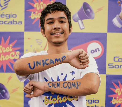 Estudante sorridente em frente a um painel institucional, usando camiseta branca, com os braços cruzados mostrando as palavras “UNICAMP” e “CIEN. COMP” escritas nos antebraços, em referência à aprovação universitária.
