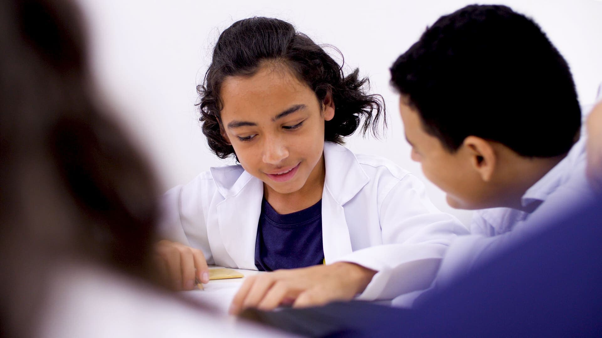 Dois estudantes usando jaleco branco realizam atividade em dupla, trocando informações enquanto observam o material sobre a mesa.