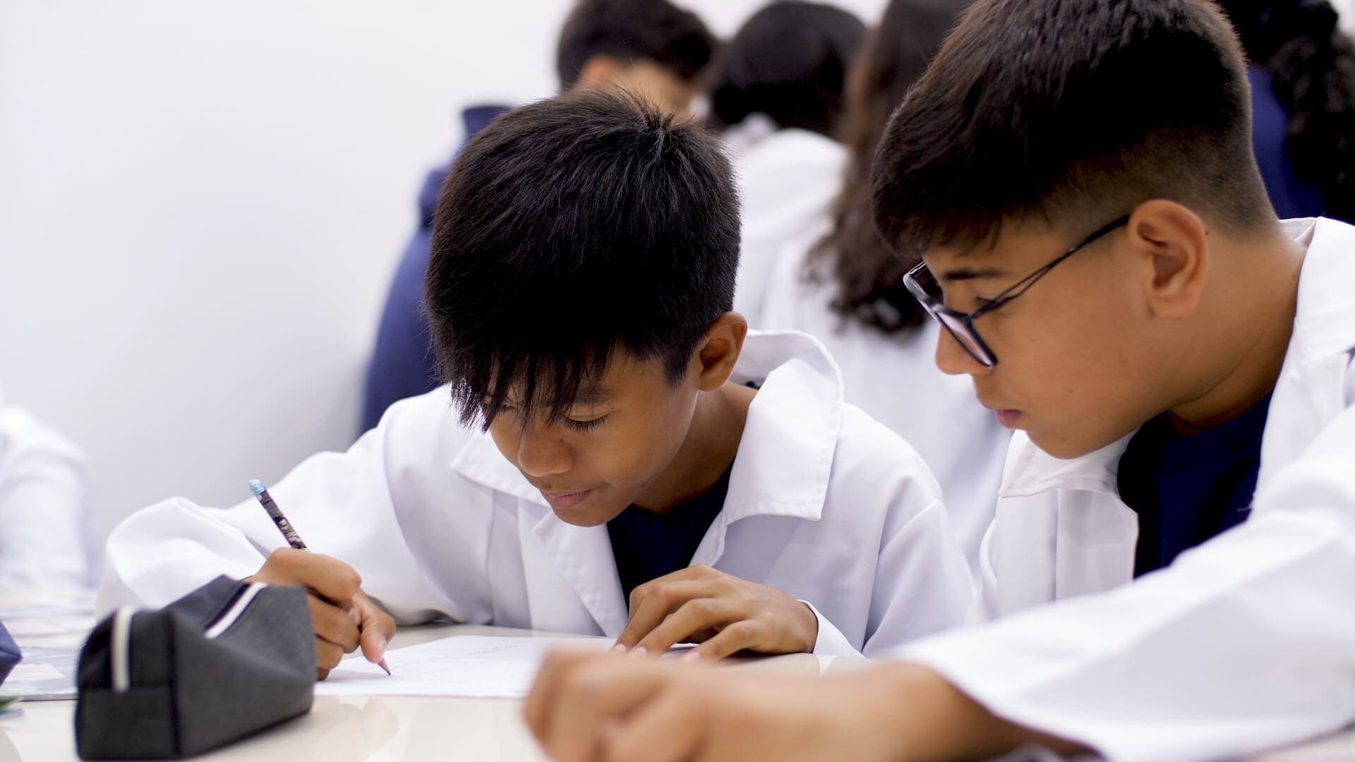 Dois estudantes usando jaleco branco escrevem em folhas sobre a mesa, concentrados em uma atividade escolar em grupo.