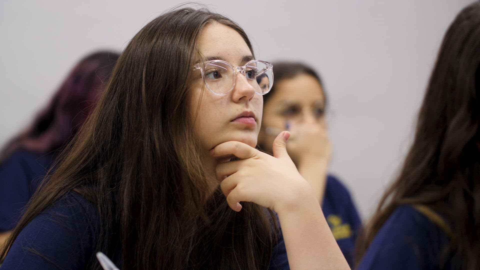 Estudante adolescente em sala de aula, usando óculos, com a mão apoiada no queixo, atenta à explicação enquanto outros alunos aparecem desfocados ao fundo.