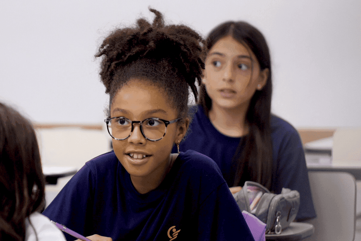Grupo de estudantes em sala de aula, usando uniforme azul, participa da atividade; uma aluna ao centro levanta a mão enquanto os colegas observam atentos.