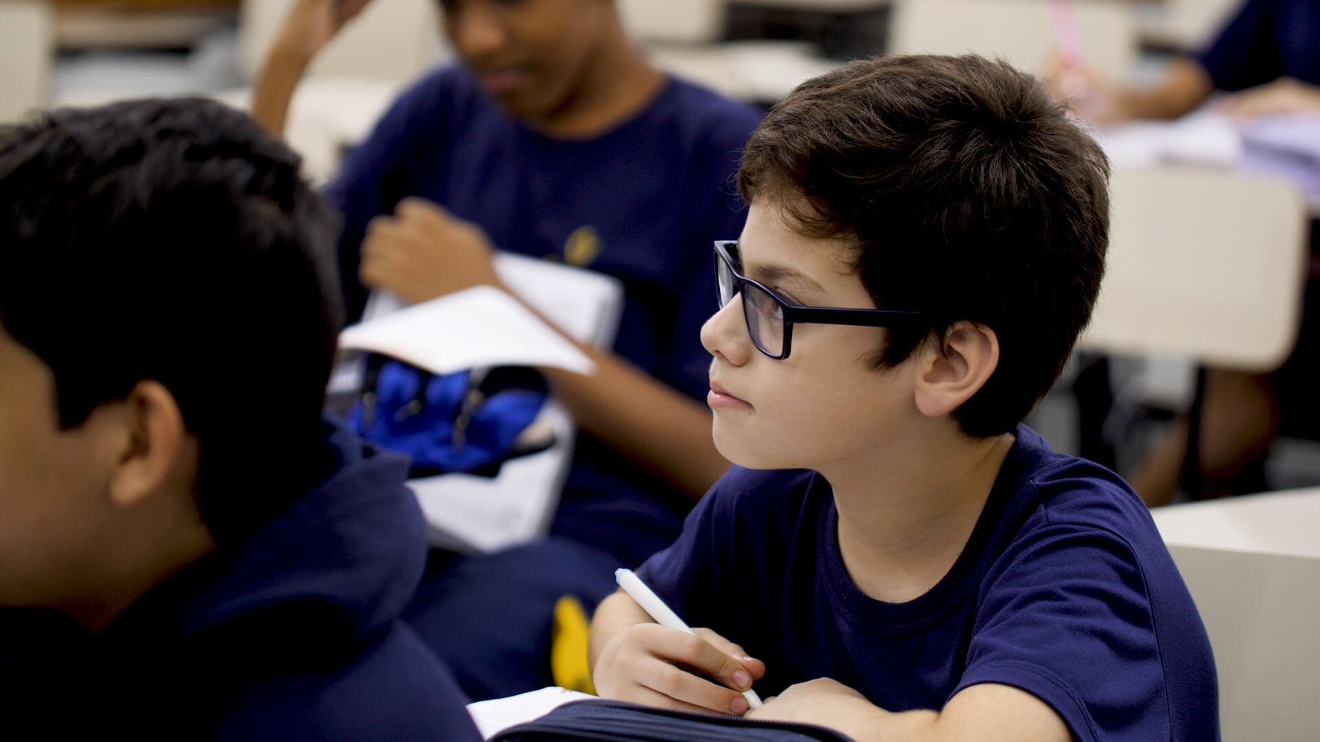 Estudante usando óculos e uniforme azul escreve em seu caderno enquanto acompanha a aula sentado em carteira escolar.