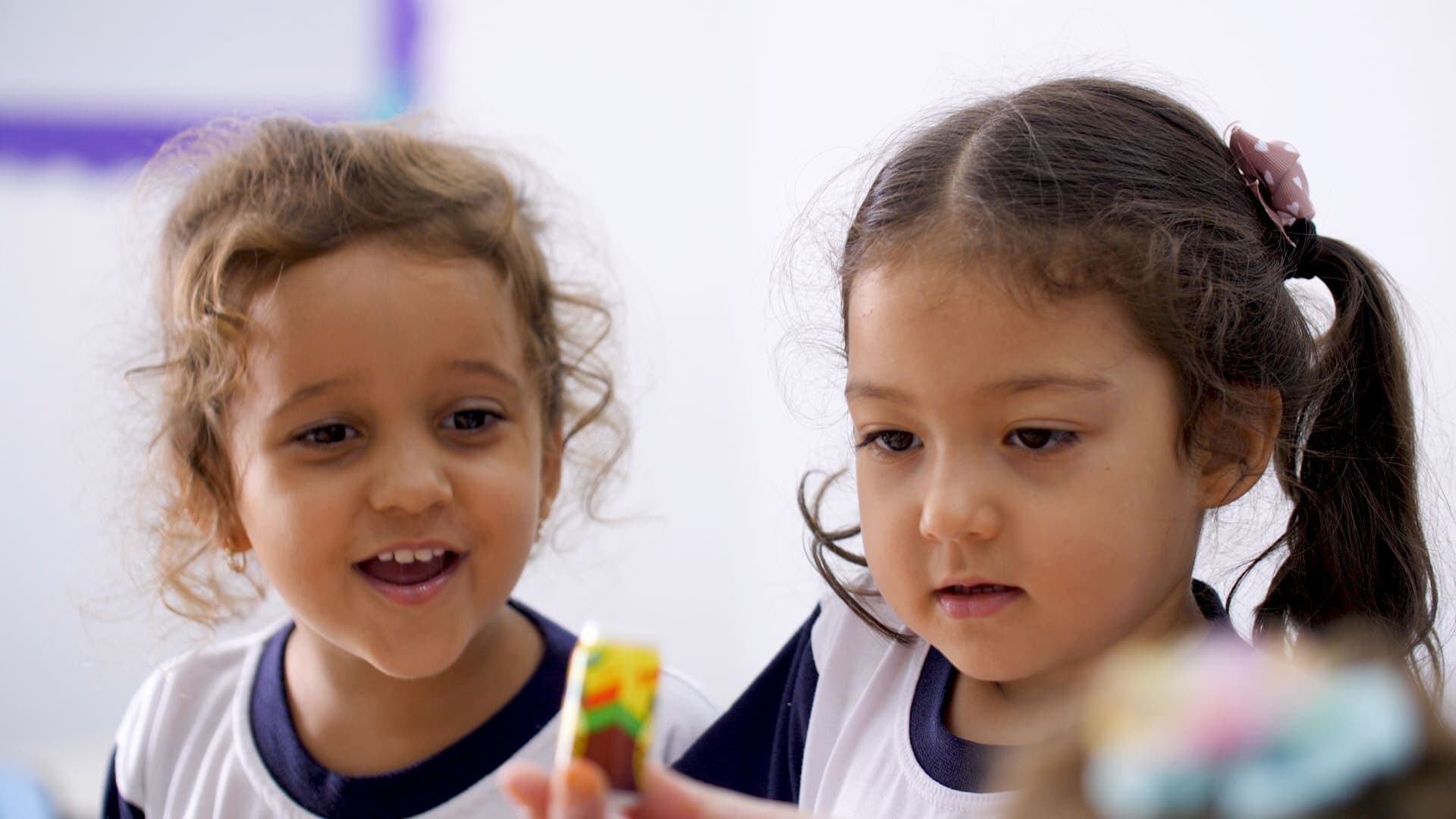 Duas crianças pequenas observam um brinquedo colorido, demonstrando curiosidade durante atividade em sala de aula.