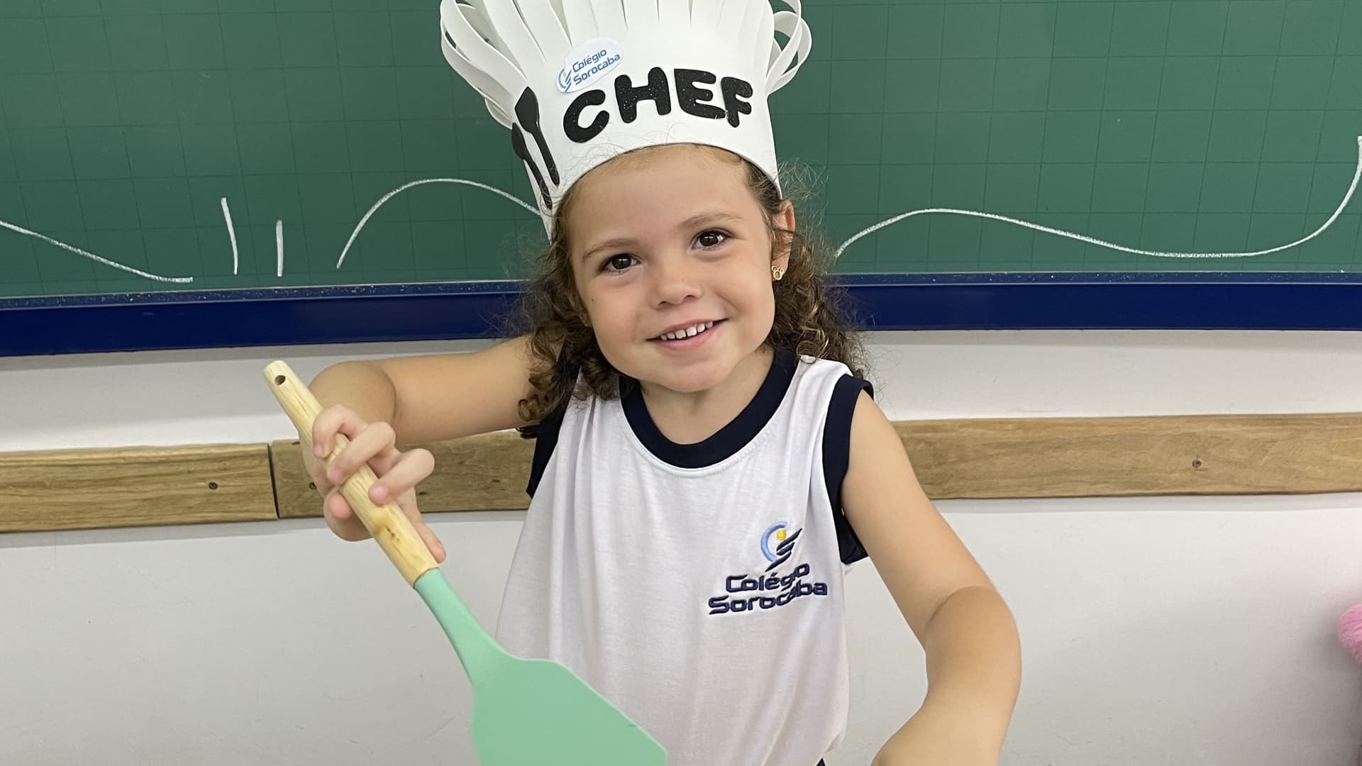 Criança sorridente usa chapéu de chef e segura uma espátula de brinquedo durante atividade lúdica em sala de aula.