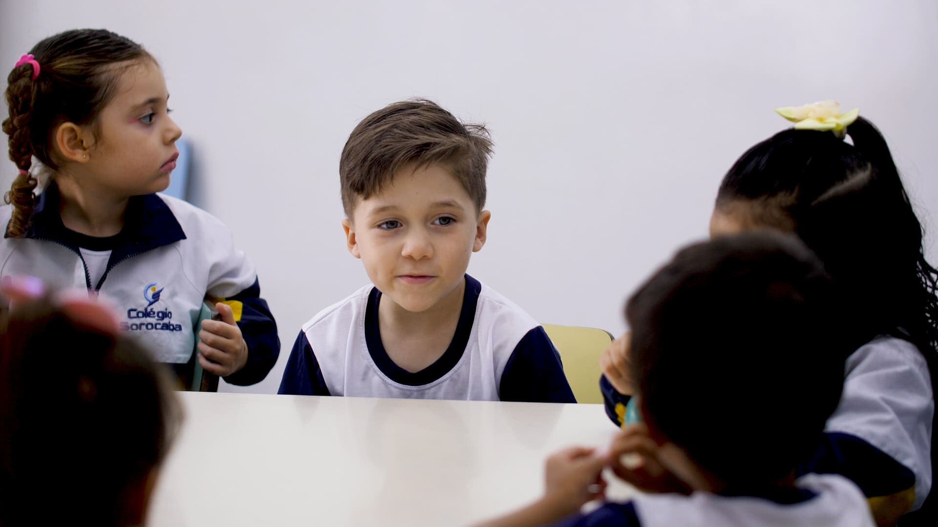 Crianças sentadas ao redor de uma mesa participam de atividade coletiva em sala de aula, interagindo entre si.