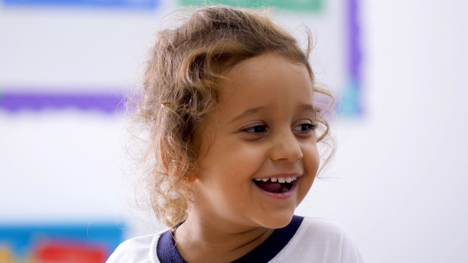 Criança pequena sorri espontaneamente em ambiente escolar, usando uniforme da escola.
