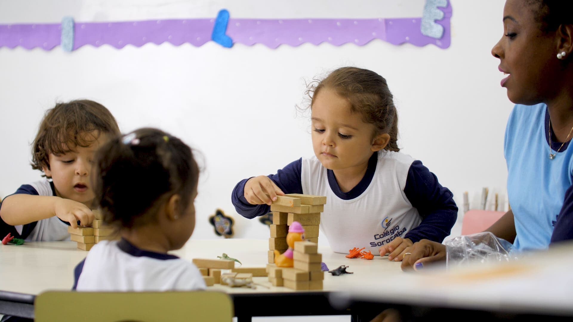 Crianças pequenas sentadas à mesa montam uma torre com blocos de madeira, acompanhadas por uma educadora em sala de aula.