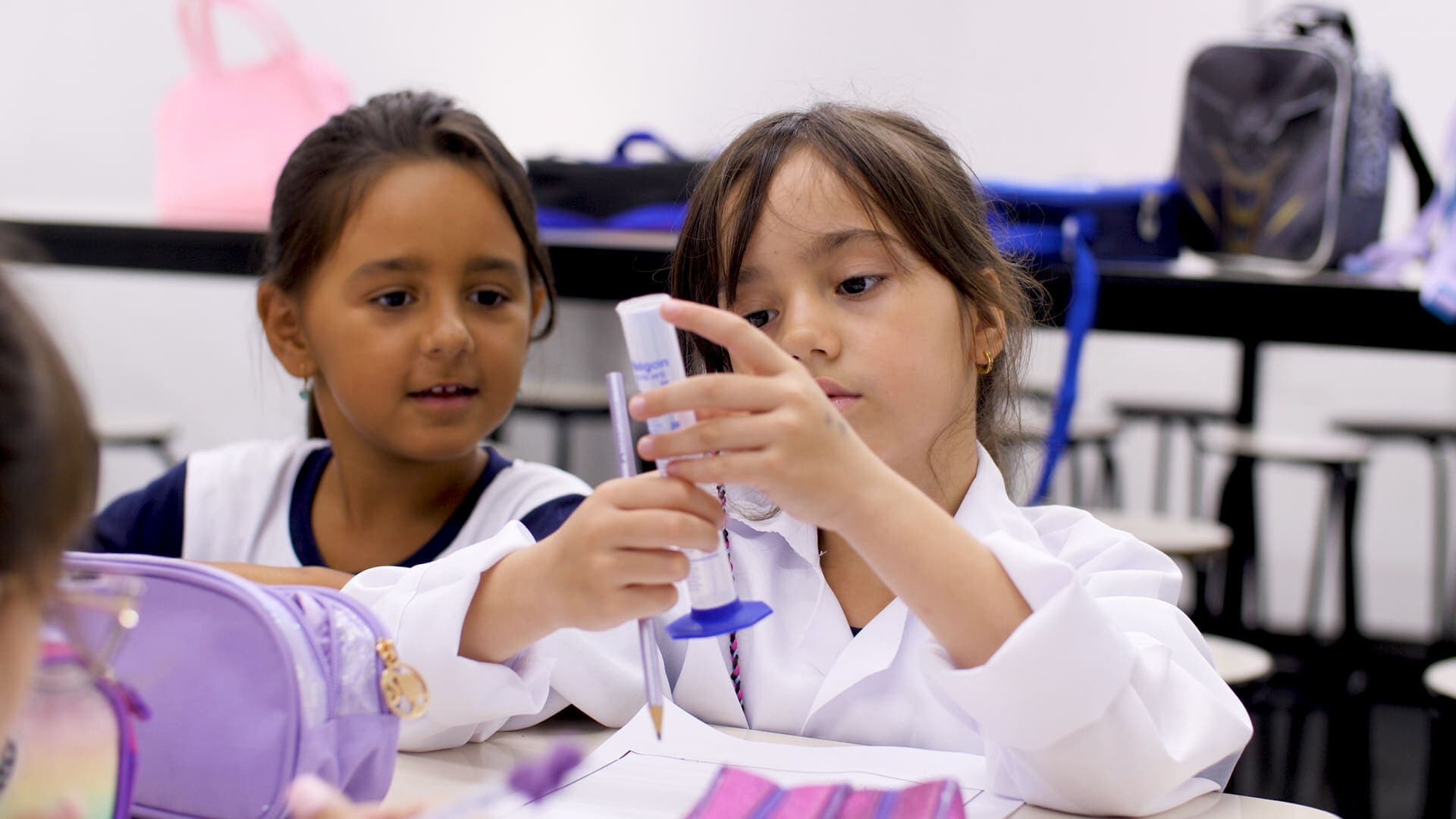 Duas estudantes em sala de aula realizam experimento com material escolar, observando atentamente o objeto que seguram juntas.