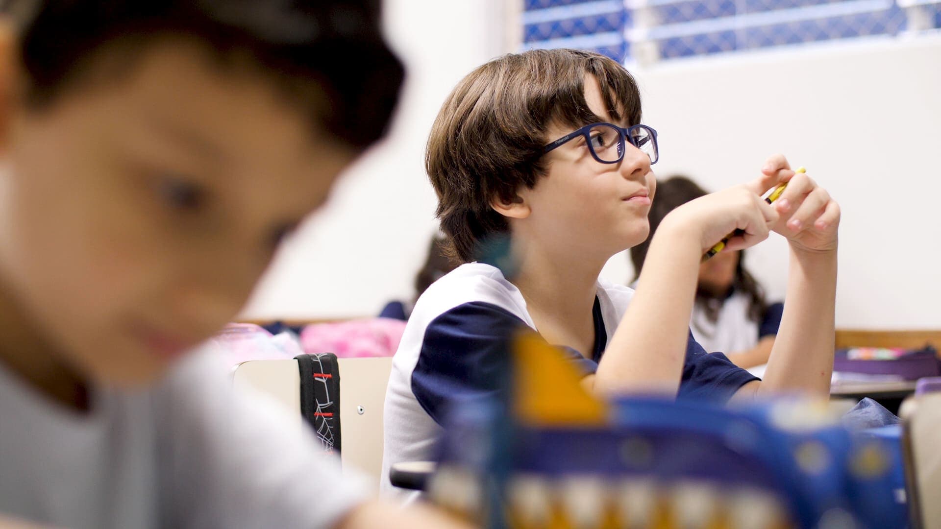 Estudante usando óculos observa a aula com atenção, sentado à carteira, com materiais escolares sobre a mesa.