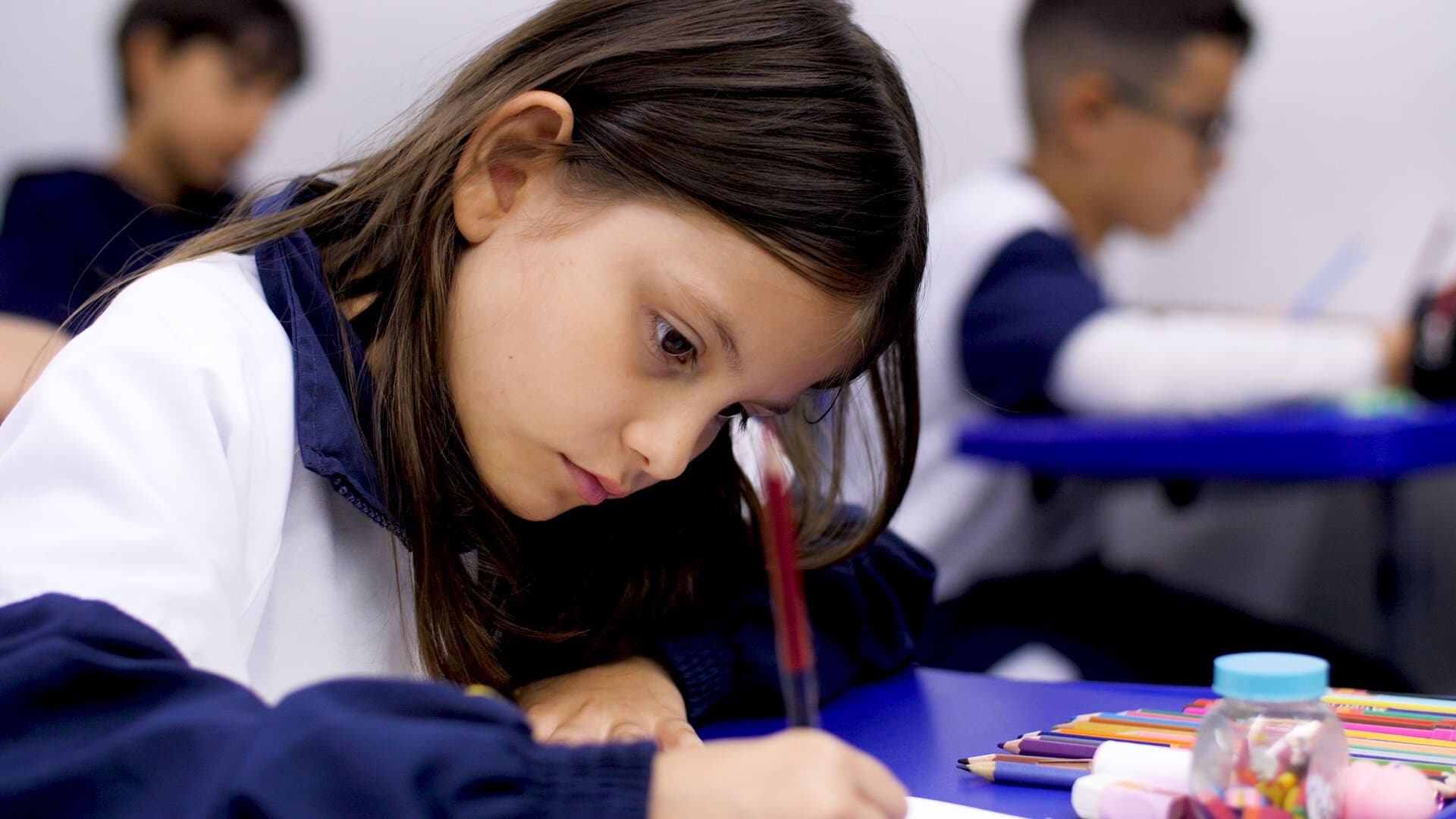 Estudante em sala de aula concentrada em atividade escrita, usando lápis sobre a mesa, com materiais escolares coloridos ao lado e outros alunos desfocados ao fundo.