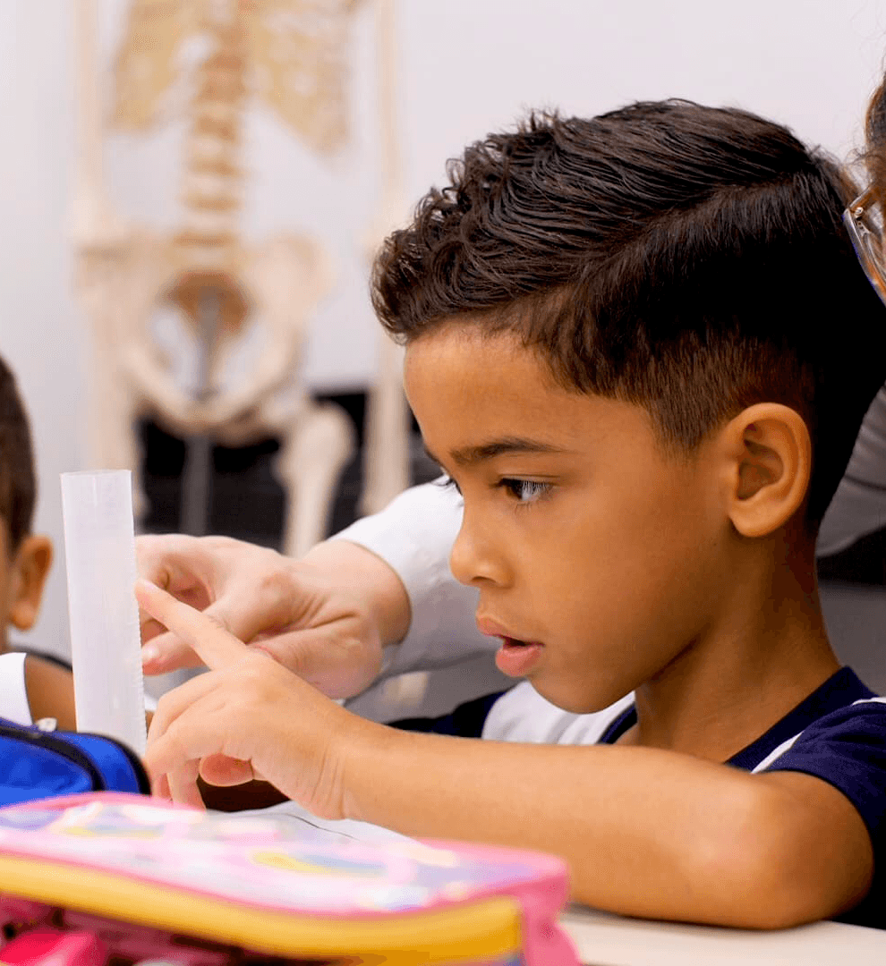 Professora orienta estudante durante atividade em sala de aula, enquanto outro aluno observa atentamente sentado à carteira.