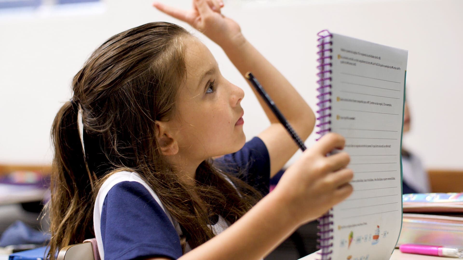 Estudante levanta a mão enquanto segura um caderno aberto, demonstrando participação ativa durante a aula.