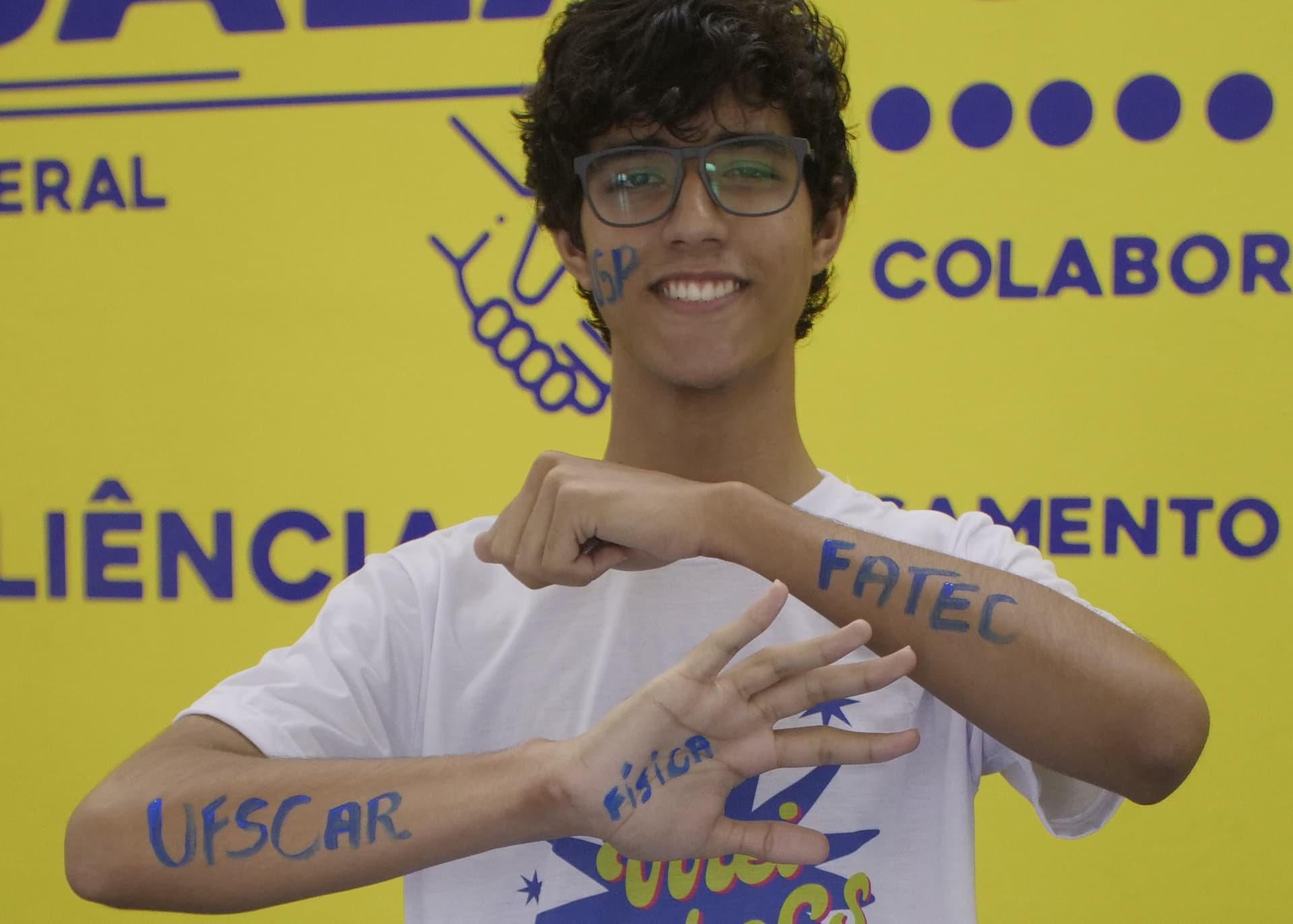 Estudante sorridente, usando óculos e camiseta branca, exibe os braços com as palavras “UFSCAR”, “FATEC” e “Física” escritas à mão, em um fundo amarelo com elementos gráficos institucionais.