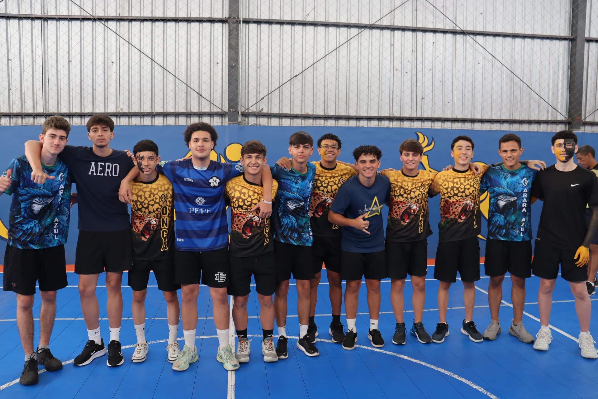 Grupo de 12 jovens atletas posando juntos em uma quadra coberta, com os braços nos ombros uns dos outros, usando uniformes esportivos azuis e amarelos com estampa de águia e onça.