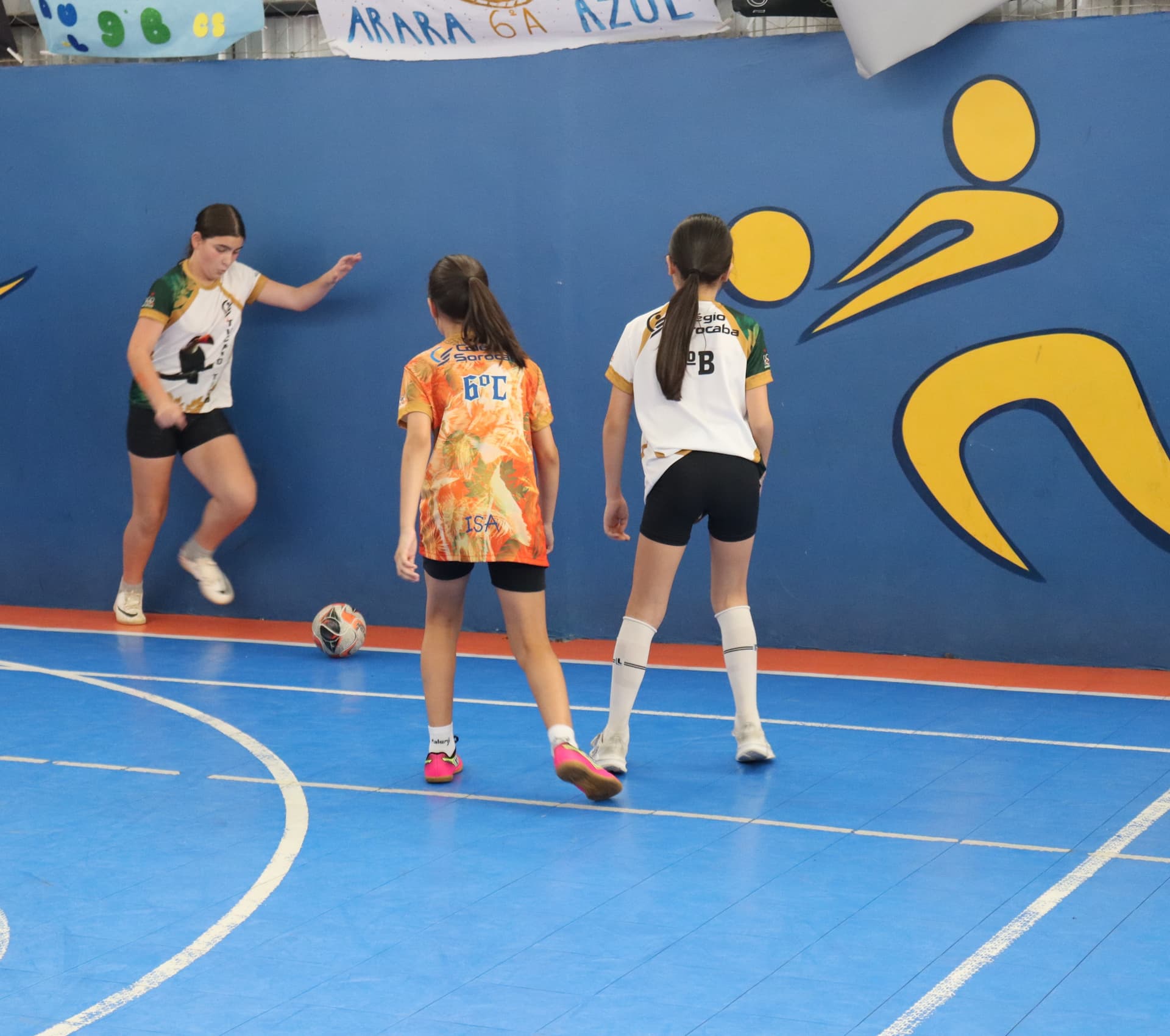 Três meninas jogam futsal em uma quadra coberta; uma delas domina a bola perto da linha lateral enquanto as outras duas se posicionam para marcar, com um grande mural azul e amarelo ao fundo.