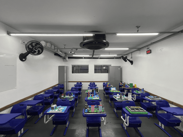 Sala de aula com carteiras individuais azuis organizadas em fileiras, trabalhos escolares expostos sobre as mesas, armários ao fundo e iluminação em LED no teto.