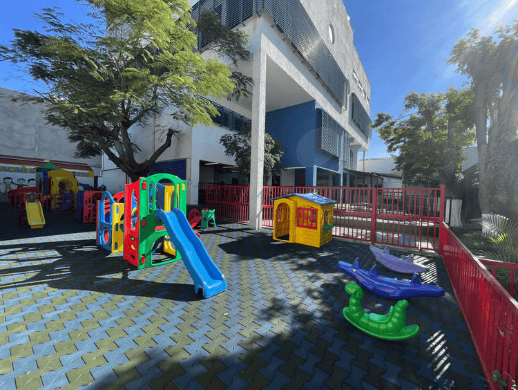 Área de playground da educação infantil com brinquedos coloridos, como escorregador, gangorras e casinha, em espaço externo arborizado e cercado.