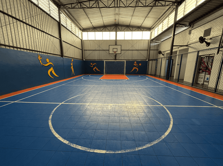 Quadra esportiva coberta do colégio, com piso azul e marcações brancas, traves de futsal, cesta de basquete e ilustrações esportivas nas paredes laterais.