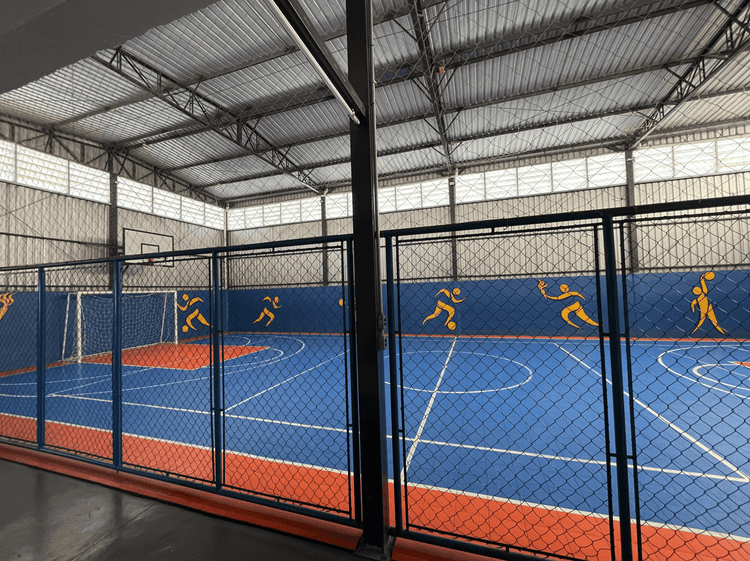 Quadra esportiva coberta com piso azul e marcações para esportes, cercada por tela de proteção, com ilustrações de atletas nas paredes laterais.