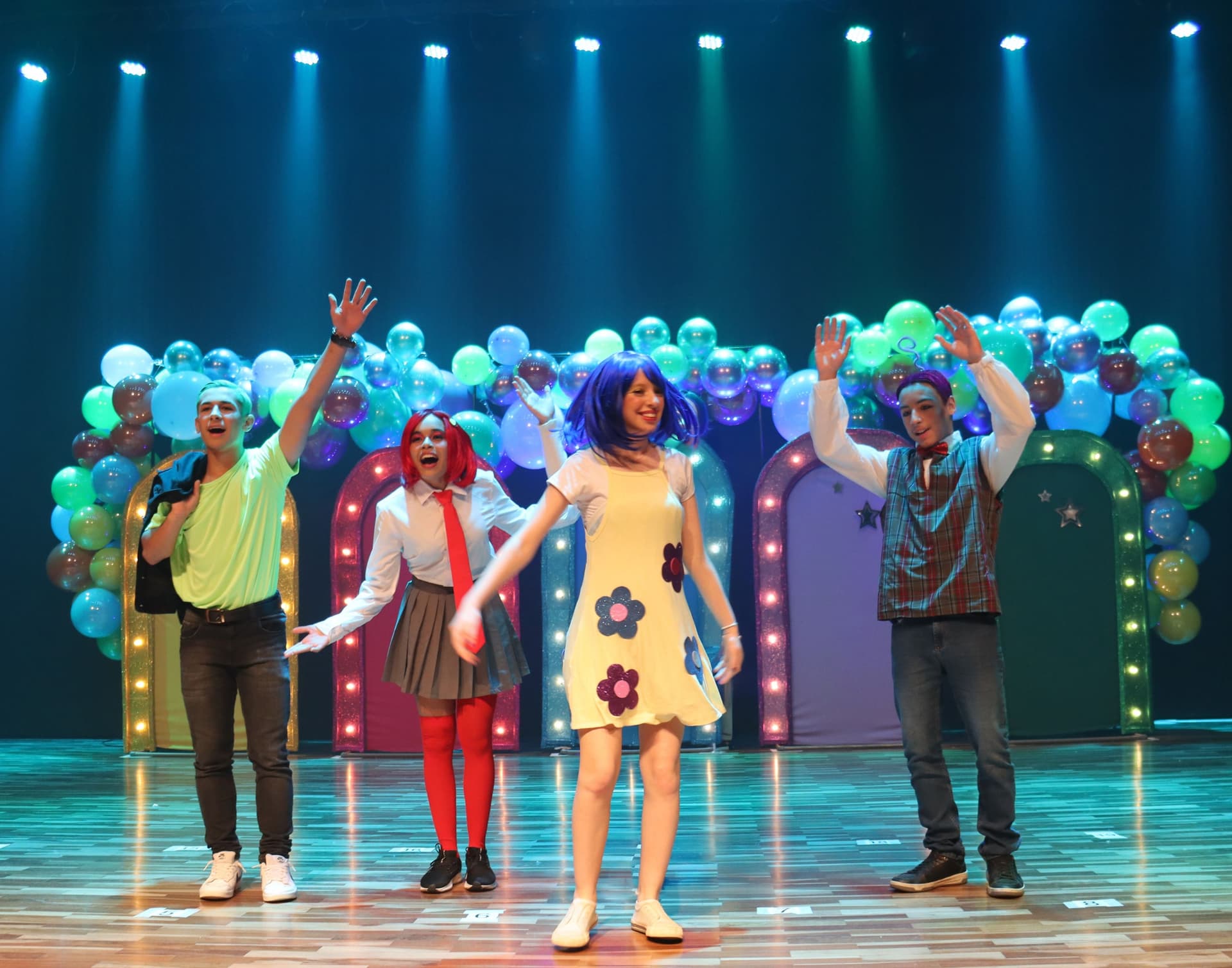 Grupo de cinco jovens em apresentação teatral sobre um palco, usando figurinos coloridos e perucas, com cenário decorado por arcos iluminados e balões ao fundo.